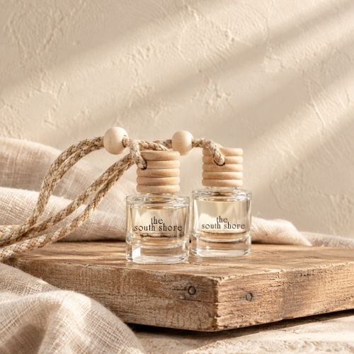 Two small clear glass reed diffuser bottles with natural wood tops and rope handles, labeled 'the dunes', displayed on a rustic wooden board with draped linen and a textured cream wall in the background.