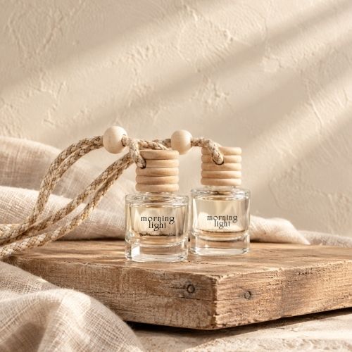 Two small clear glass reed diffuser bottles with natural wood tops and rope handles, labeled 'the dunes', displayed on a rustic wooden board with draped linen and a textured cream wall in the background.