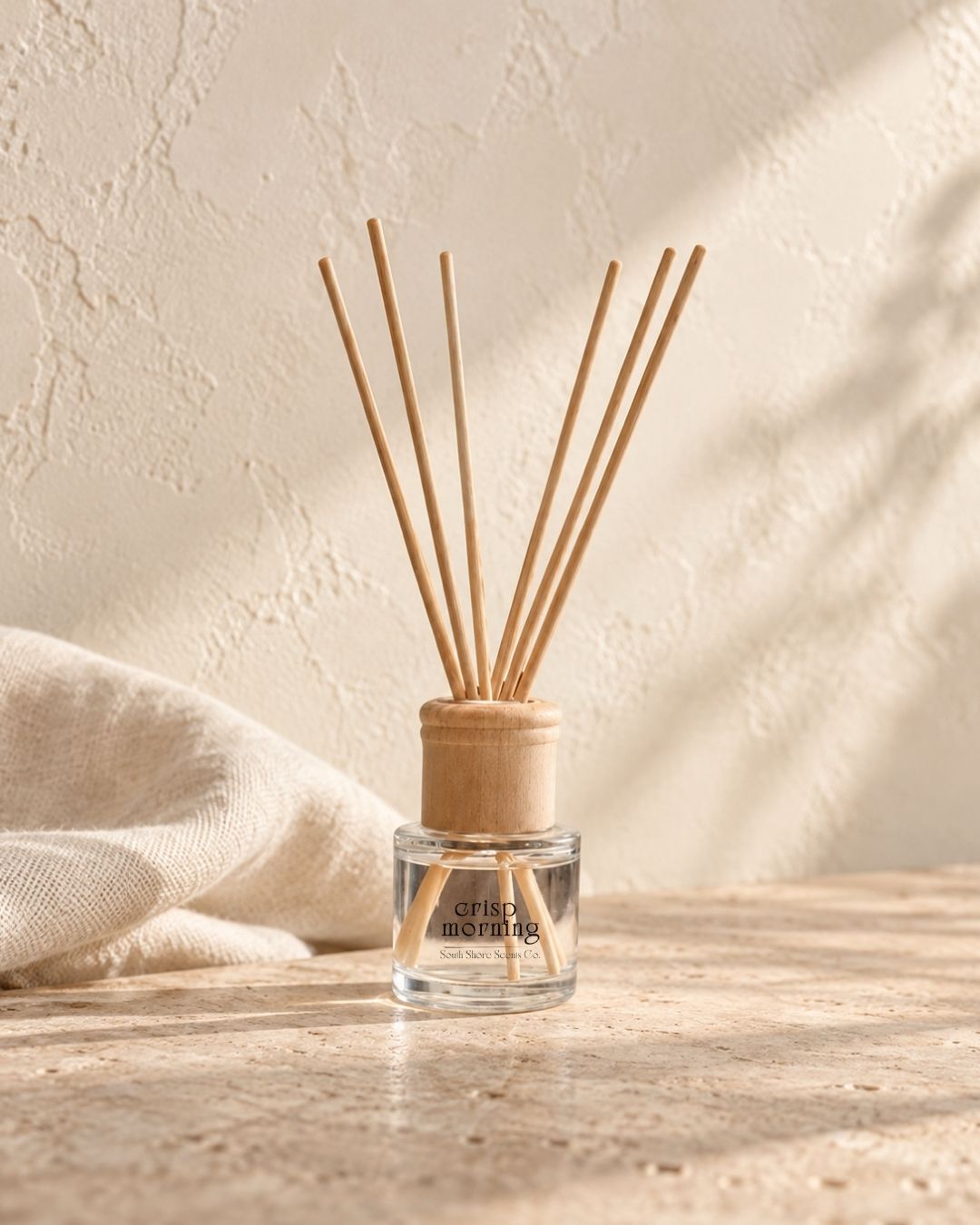 A reed diffuser bottle with a clear glass base, light wood cap, and natural bamboo reeds, placed on a stone surface with a draped cream textile and textured wall in the background.